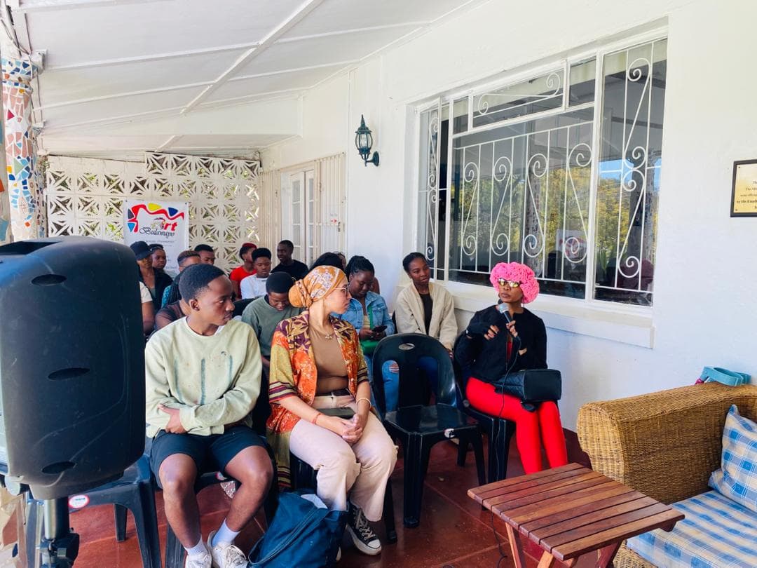 Audience listening to a woman in a pink hat speaking at an Art Bulawayo event.