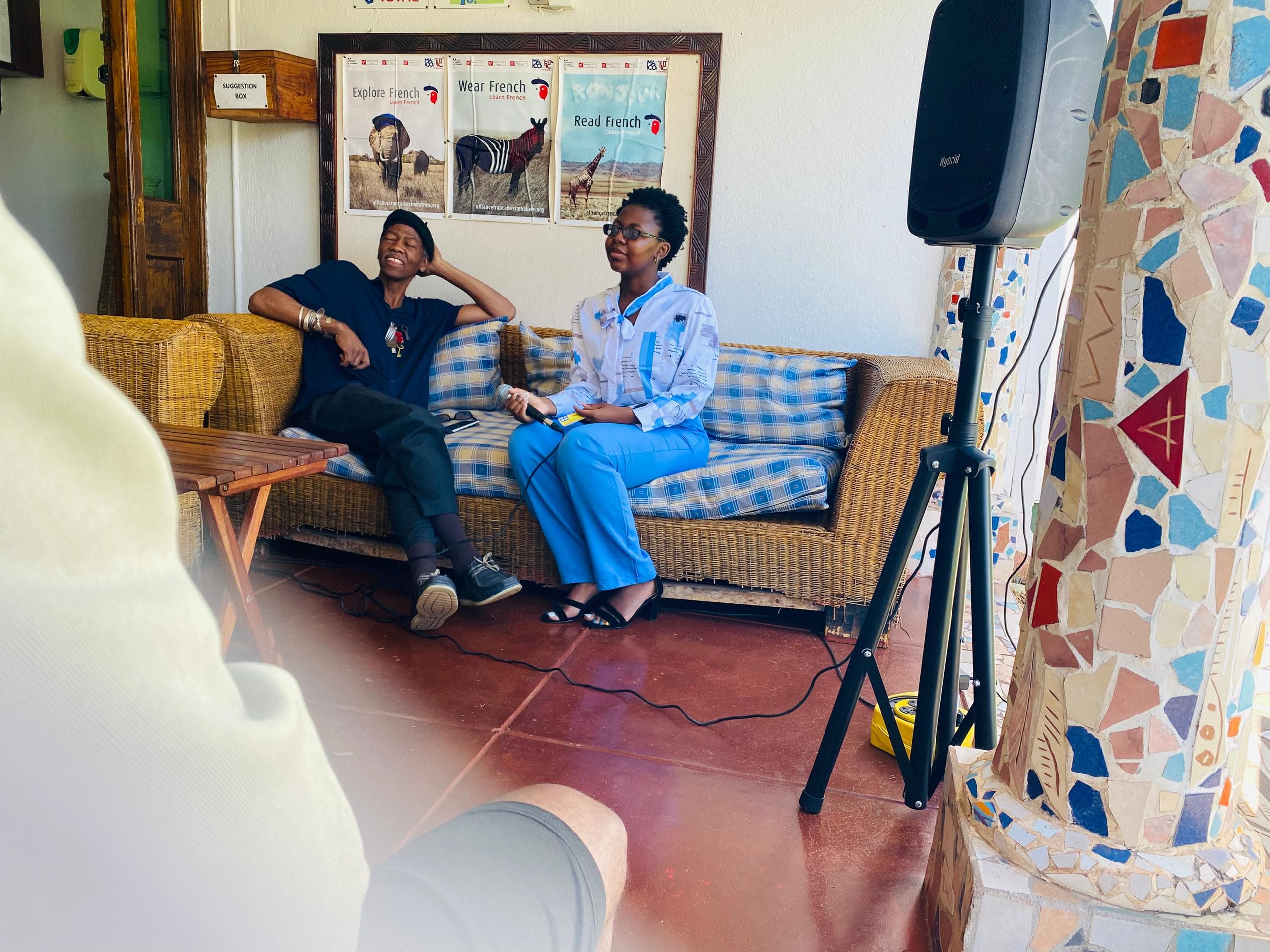 Woman interviews a person on a wicker sofa with French posters and a speaker.
